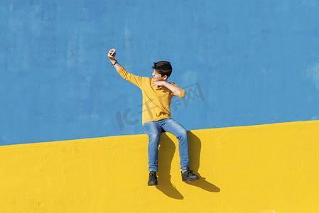 一个穿着休闲服的小男孩坐在蓝色墙壁上的黄色栅栏上，同时用智能手机自拍的前视图