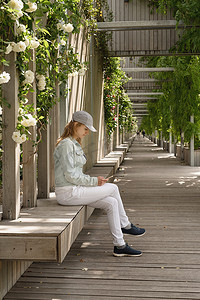 绿色公园小巷里坐在木凳上用电话的女人，墙上挂着白玫瑰