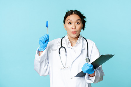 兴奋的亚洲女医生拿着剪贴板，举起笔在尤里卡标志上，有启示，提出一个想法，站在蓝色背景上