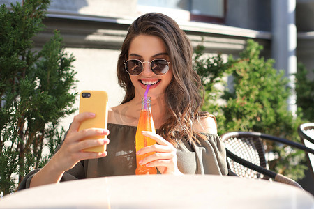 微笑的美丽黑发女人坐在街头夏季咖啡馆，用手机自拍，喝果汁。