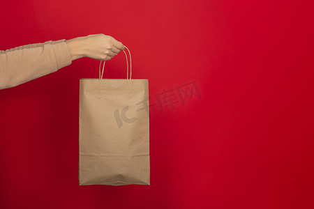 女性手握红色背景棕色牛皮纸制成的大礼品袋