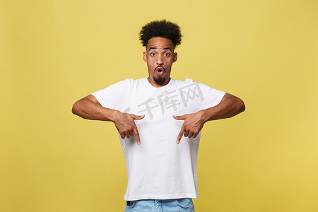 穿着休闲白衬衫的年轻非洲裔美国男子惊讶不已，他的表情兴奋迷人，用食指指着金黄色背景上的复制空间，为您的文字或促销内容