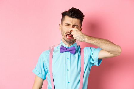 照片中，一名男子因恶心的气味而哭泣，闭上鼻子以免发出难闻的臭味，站在粉色背景的领结和吊带上