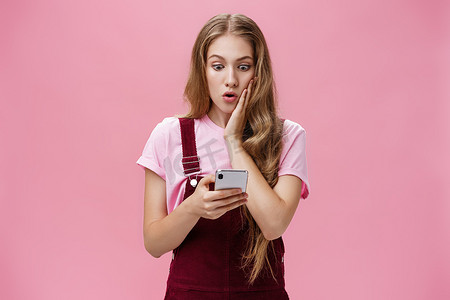 发现了摄影照片_女孩在阅读智能手机中的信息后发现了令人震惊的事实，她惊讶地抱住脸颊，关切地盯着手机屏幕，对粉红色墙壁上的意外消息做出反应