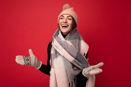 照片中，美丽、快乐、有趣、年轻的黑发女人被隔离在红色背景墙上，戴着冬季围巾手套和温暖的帽子，看着旁边，玩得很开心