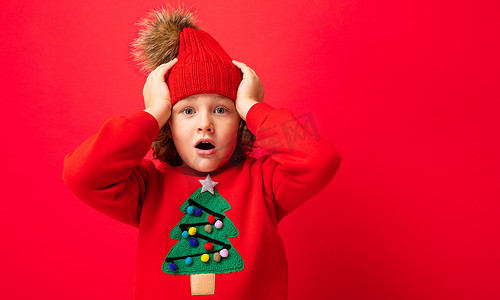 戴着温暖帽子和毛衣、红色背景圣诞树的酷金发小孩在闲逛，圣诞节概念