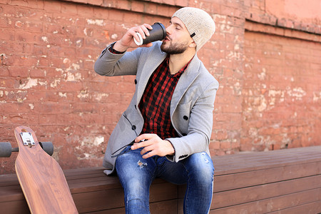 穿着灰色外套和帽子的英俊年轻人，休息，坐在长板旁喝咖啡。