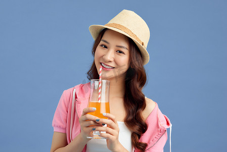 快乐的年轻亚洲女性戴着草帽，在蓝色背景下喝果汁的肖像。
