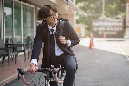 身穿西装的英俊年轻亚洲商人早上骑着自行车微笑着去上班。软焦点