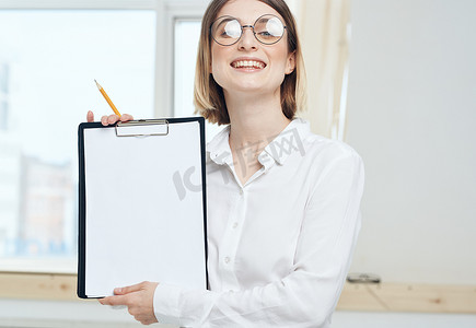 一位女士拿着一个文件夹，里面有一张白色的样机纸，背景是一扇窗户