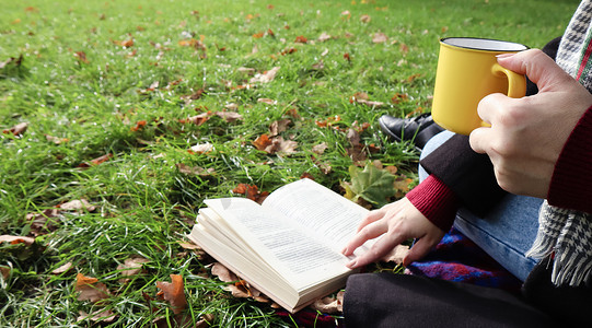 在温暖的一天，在绿色的草地上，一个女孩坐在秋天森林的毯子上，看书，喝着城市公园里黄色杯子里的热饮。