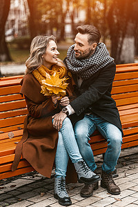 一对相爱的情侣坐在公园的长凳上，穿着外套和围巾，拥抱着一束落叶。