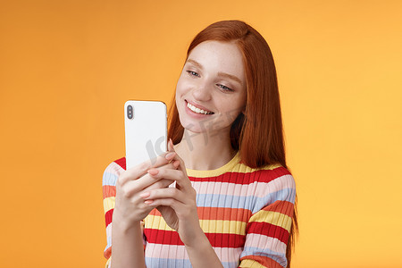 可爱的女性柔嫩年轻红发魅力女孩拿着智能手机拍照夏季都市氛围女博主拍摄帖子在线故事快乐地站着橙色背景微笑