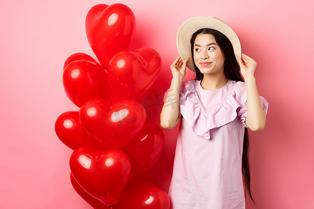 温柔浪漫的亚洲少女在约会时戴着草帽和裙子，站在情人节心形气球附近，带着梦幻般的微笑、粉色背景望向一边