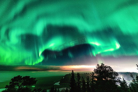 瑞典北部，斯堪的纳维亚半岛，灿烂的绿色极光在山区的瑞典雾蒙蒙的森林景观中闪耀，村庄的光线和北极光以不同柔和的色彩为天空着色