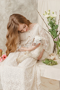 抱着蛋糕的女孩摄影照片_这个女孩坐在复活节餐桌旁，手里拿着蛋糕、春天的花朵和鹌鹑蛋，怀里抱着一个白色的孩子。