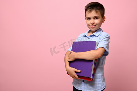 可爱的男学生手里拿着书，在镜头前摆出可爱的微笑，孤立在粉红色的背景上，有文字空间。