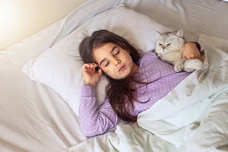 猫睡觉摄影照片_一个穿着紫色睡衣的黑发小女孩抱着一只白色迷人的猫睡觉，它躺在床上的白色枕头上。