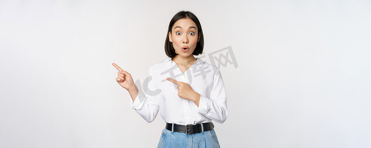 兴奋的年轻亚洲女性的肖像，办公室女士指着打折的手指，展示销售横幅，站在白色背景上