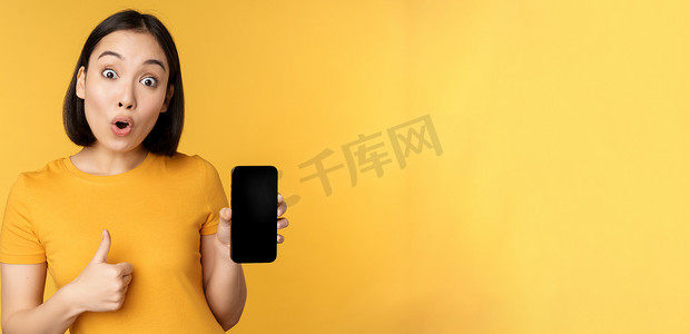 兴奋的亚洲女性展示手机屏幕，竖起大拇指，喜欢某事，推荐智能手机应用程序，站在黄色背景上