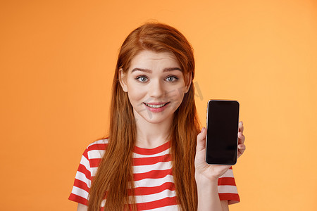 特写可爱的白种红发女孩显示智能手机屏幕橙色背景，微笑可爱，推荐应用程序，推广应用程序，在线优惠销售，介绍手机