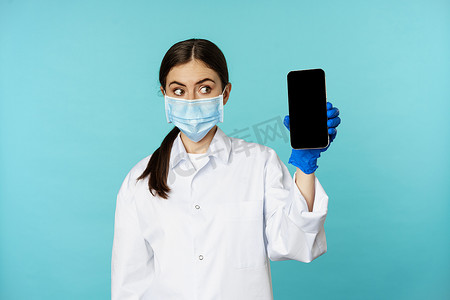 手机界面展示摄影照片_身穿医用面罩和医院制服的年轻女医生，展示手机应用程序、屏幕界面、在线诊所概念，站在绿松石背景上