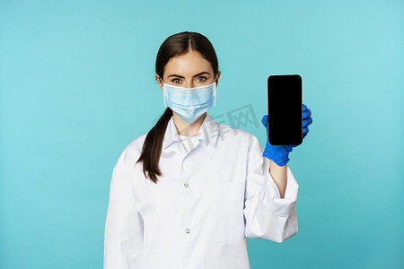 小程序界面摄影照片_身穿医用面罩和医院制服的年轻女医生，展示手机应用程序、屏幕界面、在线诊所概念，站在绿松石背景上