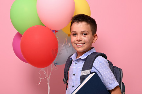 背着背包、拿着书和彩色气球的男学生，在粉红色的背景下对着镜头可爱地微笑着，并有复印空间。