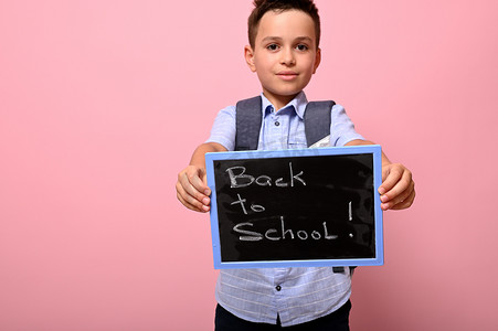 背着背包的可爱男学生拿着写有粉笔字样的黑板，回到学校。