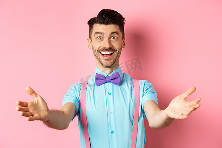 热烈欢迎摄影照片_戴领结的快乐帅哥伸出双手热烈欢迎，邀请或问候您，接受礼物，站在粉红色的背景上