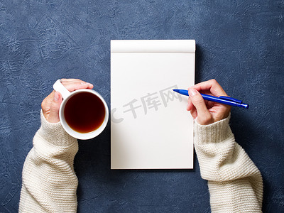女人在深蓝色桌子上的笔记本上写字，手里拿着铅笔、一杯茶、素描本绘图的衬衫