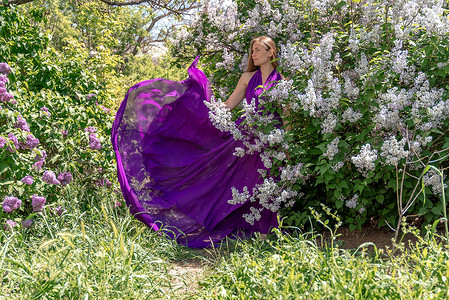 丁香花中的时装模特，穿着美丽长裙的年轻女子在风中飘扬，盛开的花园中的户外美女肖像
