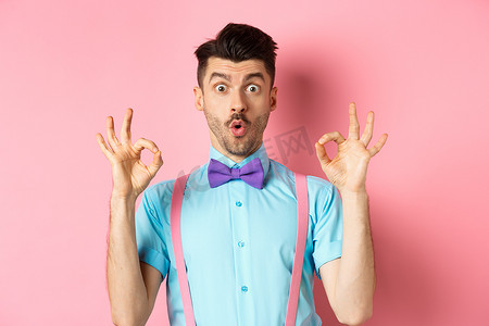 酷炫产品背景摄影照片_打领结的白人男子印象深刻，查看酷炫的报价，表现出良好的姿态，说哇，赞扬好产品，站在粉红色的背景上