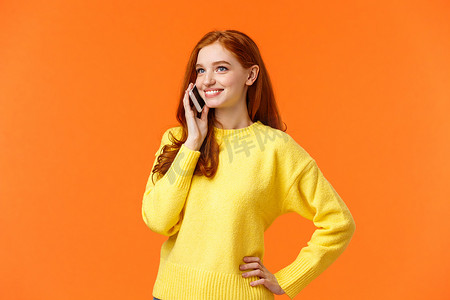 腰部肖像漂亮的红发女性打电话给朋友，交谈，在假期购物时确认在线订单，无忧无虑地站立，将智能手机放在耳边，橙色背景