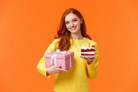 橙色生日摄影照片_可爱的红发女孩带着包装好的礼物和一块美味的生日蛋糕来参加聚会，请朋友吹灭蜡烛，与庆祝女朋友生日一起庆祝，在橙色背景中微笑