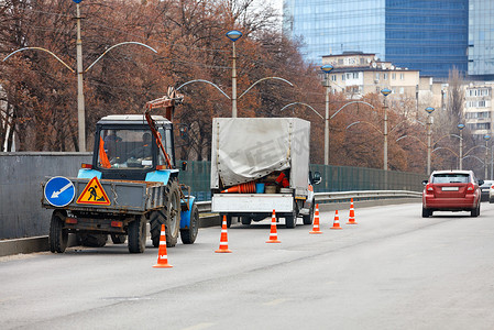 一群道路服务人员驾驶着一辆旧的公用拖拉机和一辆卡车，正在用橙色锥体封闭一段道路，以便进行后续清洁。