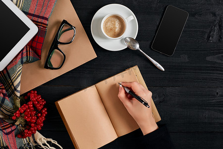 茶手写摄影照片_早上，年轻女子右手在木桌上的空白笔记本上写字，旁边放着咖啡杯、智能手机和眼镜