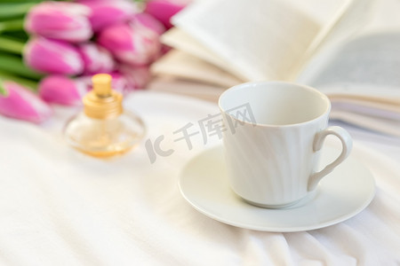 书茶摄影照片_春天静物画，配有白色杯子、一瓶香水、一本打开的书和一束美丽的粉红色郁金香