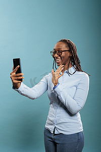 美丽自信的年轻女士站在蓝色背景上通过在线电话视频通话问候朋友。