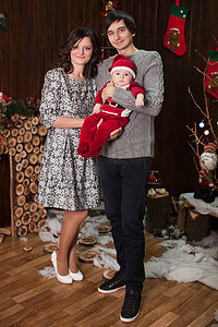 小孩圣诞老人摄影照片_一家人带着一个穿着圣诞老人服装的小孩在家里的壁炉旁。
