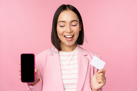 身穿西装的微笑韩国女商人，展示手机屏幕、信用卡、展示网上银行应用界面、粉色背景