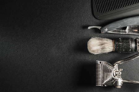 黑色表面上是旧理发工具。两个老式手动理发器、梳子、剃刀、剃须刷、美发剪刀。
