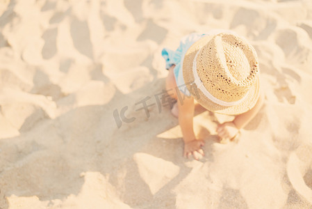 夏天，戴着草帽、穿着蓝色裙子的女婴在海滩上玩沙子。