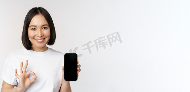手机批准摄影照片_微笑的亚洲女性表现得不错，手机屏幕，推荐智能手机应用程序，站在白色背景上