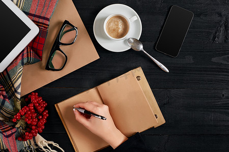 早上，年轻女子右手在木桌上的空白笔记本上写字，旁边放着咖啡杯、智能手机和眼镜