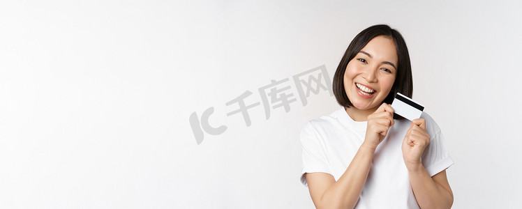 微笑的亚洲女性拥抱信用卡、购买非接触式产品、站在白色背景上的白色 T 恤的形象