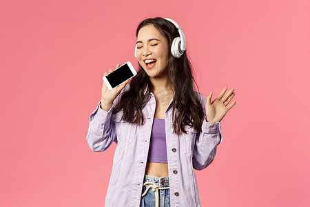 时尚快乐女人的腰部肖像，使用耳机聆听精彩的新曲目，下载卡拉 OK 应用程序并使用智能手机（如麦克风），唱着精彩的音乐