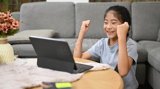 快乐的亚洲孩子坐在客厅里，通过电脑平板电脑在虚拟课堂上在线学习。
