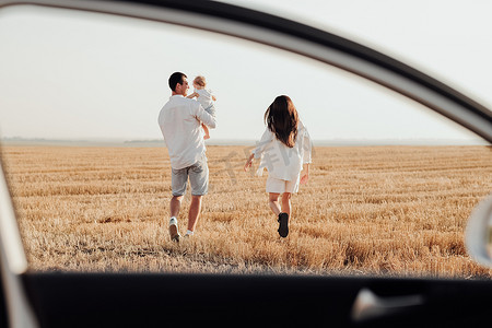 孩子散步摄影照片_日落时分，年轻的家庭在田野里散步，透过车窗看后景，爸爸带着女儿与妈妈共度周末