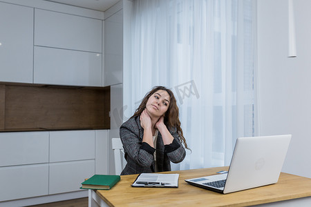 年轻漂亮的女商人坐在家里的桌子旁，在笔记本电脑上在线工作，疲惫，伸展，经历身体疼痛，处理文件